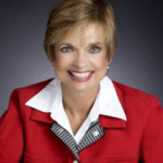 Colleen Burcar - Head-and-shoulders portrait of a smiling woman with short blond hair, wearing a red blazer over a white collared shirt.