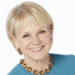 Smiling older woman with short blonde hair wearing a blue sweater and beaded necklace, facing forward against a light background
