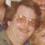 Smiling man with brown hair, mustache, and yellow-tinted glasses at a social gathering, with people in the background.
