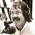 Radio host with a mustache wearing headphones, speaking into a microphone in a studio in black-and-white. (informative)