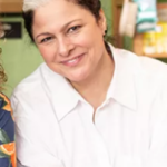Ann Delisi - Smiling woman with short gray-streaked hair wearing a white button-down shirt, seated in a shop interior.