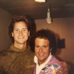 Brad Hurtado and Richard Simmons at WXYZ's Kelly & Compmany7 - Two men posing indoors, smiling at the camera; the younger man wears a brown patterned sweater, the older man wears a colorful jacket and has his arm around him.