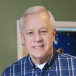 Portrait of an older man with short gray hair, wearing a blue plaid shirt, smiling at the camera in a indoors setting