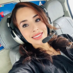 Woman wearing an aviation headset with microphone, smiling at the camera in a small aircraft cabin.