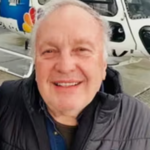 WDET: Helicopter Pilot.Smiling older man in a black puffer vest posing in front of a white helicopter on a tarmac surface.