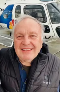 WDET: Helicopter Pilot.Smiling older man in a black puffer vest posing in front of a white helicopter on a tarmac surface.