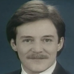 Jay Brandow - Head-and-shoulders portrait of a man with dark hair and a mustache, wearing a suit and tie, against a blue backdrop