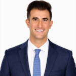 Smiling man in a navy suit, white shirt, and blue tie against a white background (professional headshot).