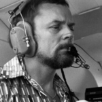 Man wearing large aviation headset with microphone, focused in a cockpit or control cabin.
