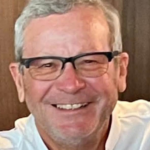 Smiling older man with gray hair and black glasses, wearing a light-colored shirt.