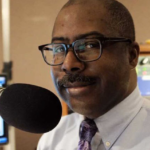 Jerome Vaughn - Portrait of a man with glasses and a mustache speaking into a large microphone in a radio studio.