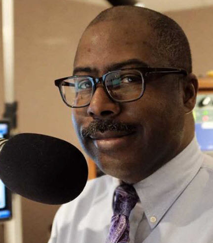 Vaughn2webp - BroadcastingVault.com Jerome Vaughn - Portrait of a man with glasses and a mustache speaking into a large microphone in a radio studio.